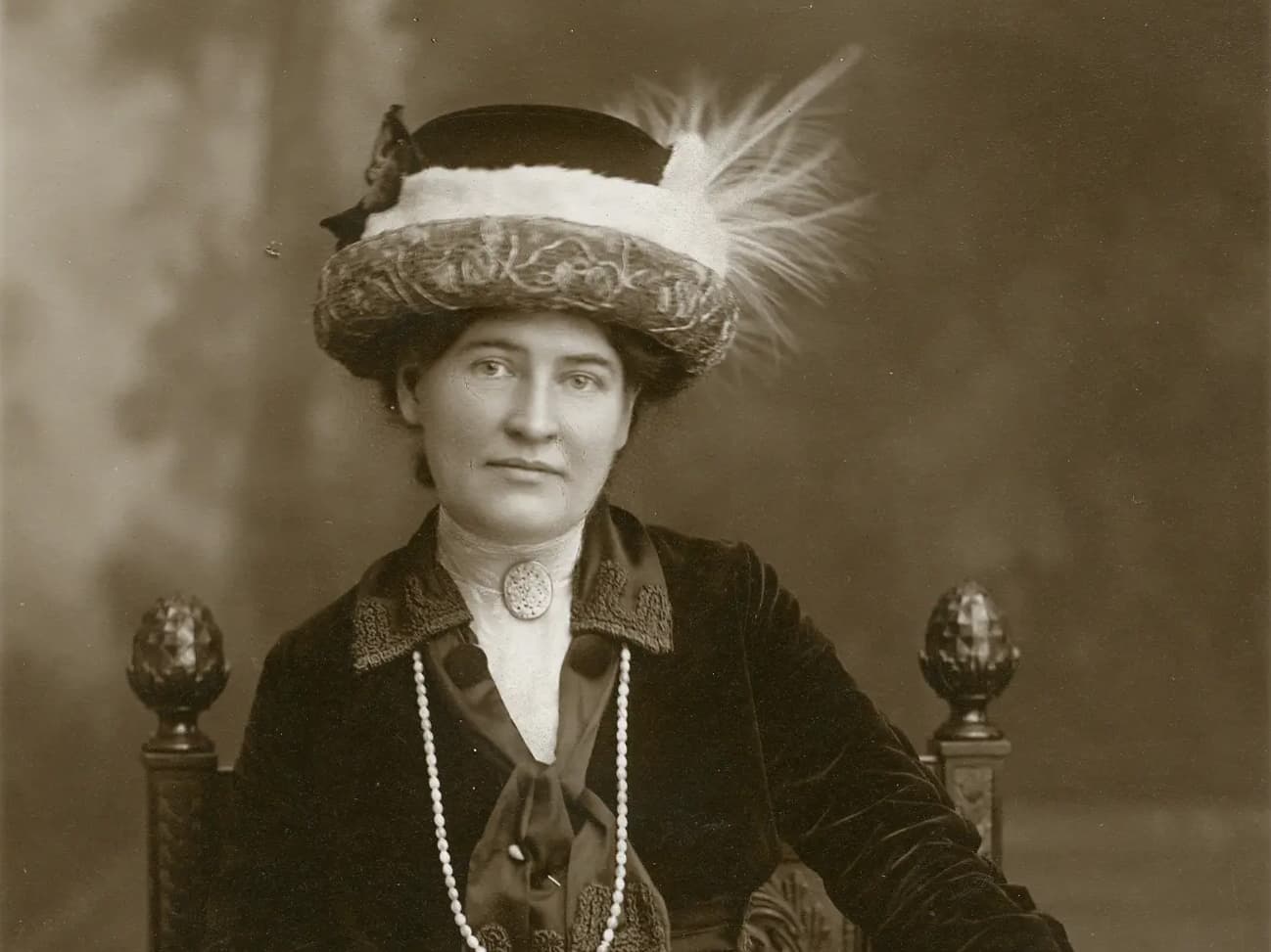 Black and white portrait of a woman seated, wearing a large feathered hat and a pearl necklace. She gazes calmly towards the camera, dressed in early 20th-century attire, against a blurred backdrop.