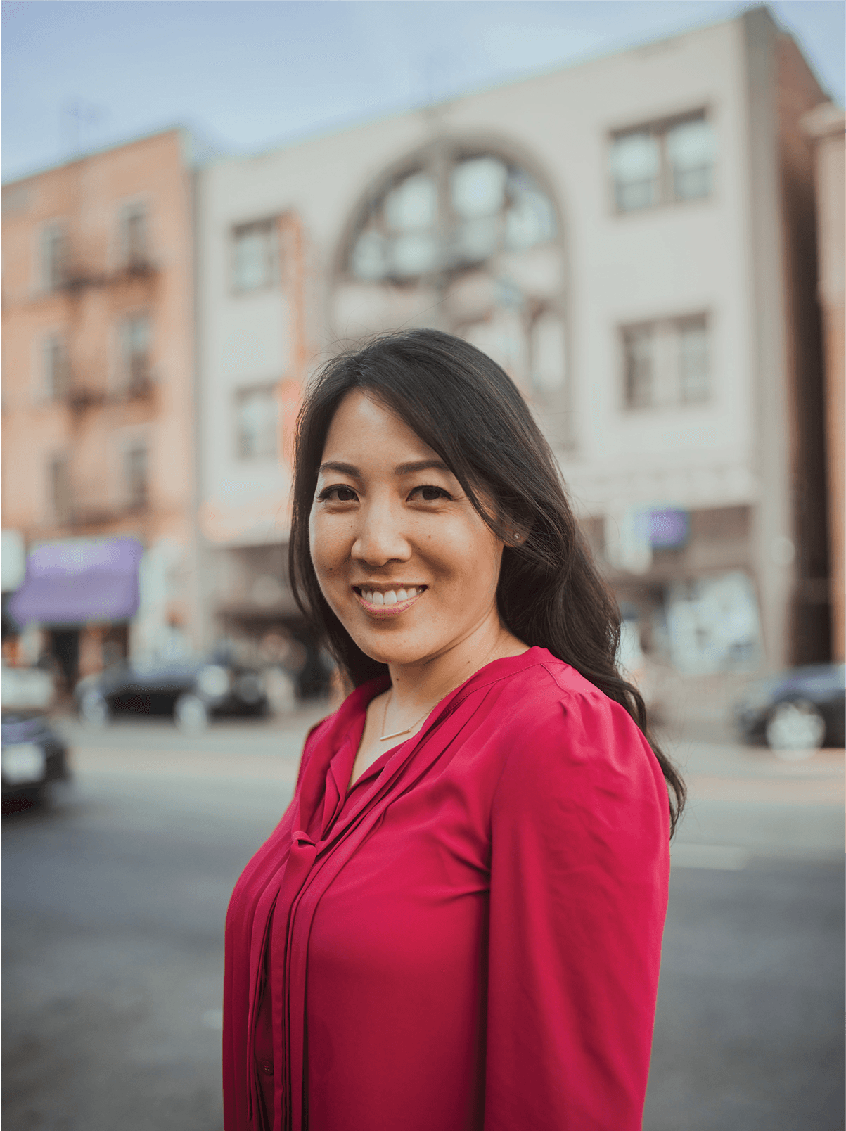 A woman with long dark hair wearing a bright pink blouse smiles while standing on a city street. The background is blurred, showing vehicles and buildings.