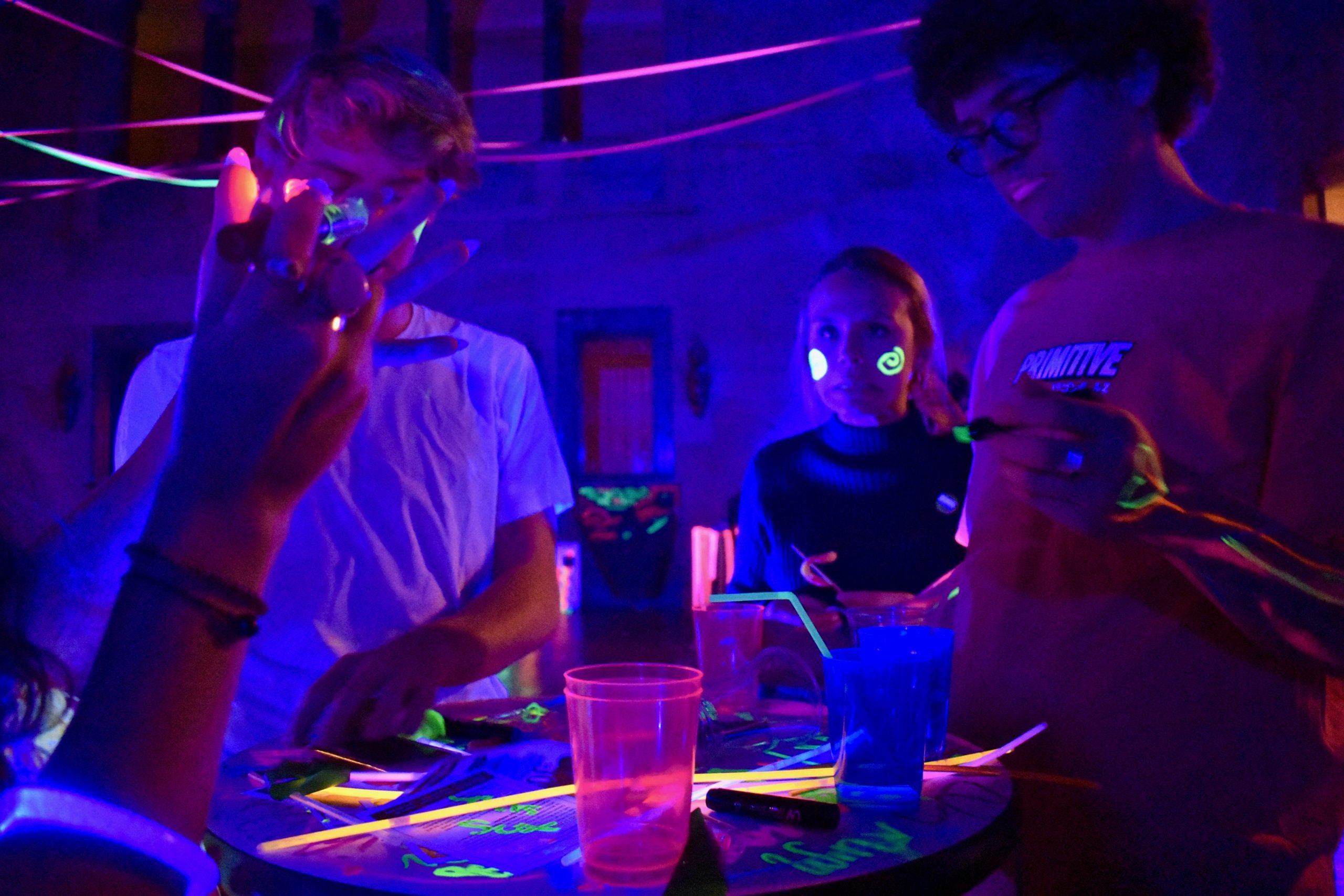 A group of people are enjoying a neon-themed party. The room is illuminated with UV lights, making their clothing and painted designs glow brightly. They are gathered around a table with neon art materials and illuminated drinks. The atmosphere is lively and colorful.