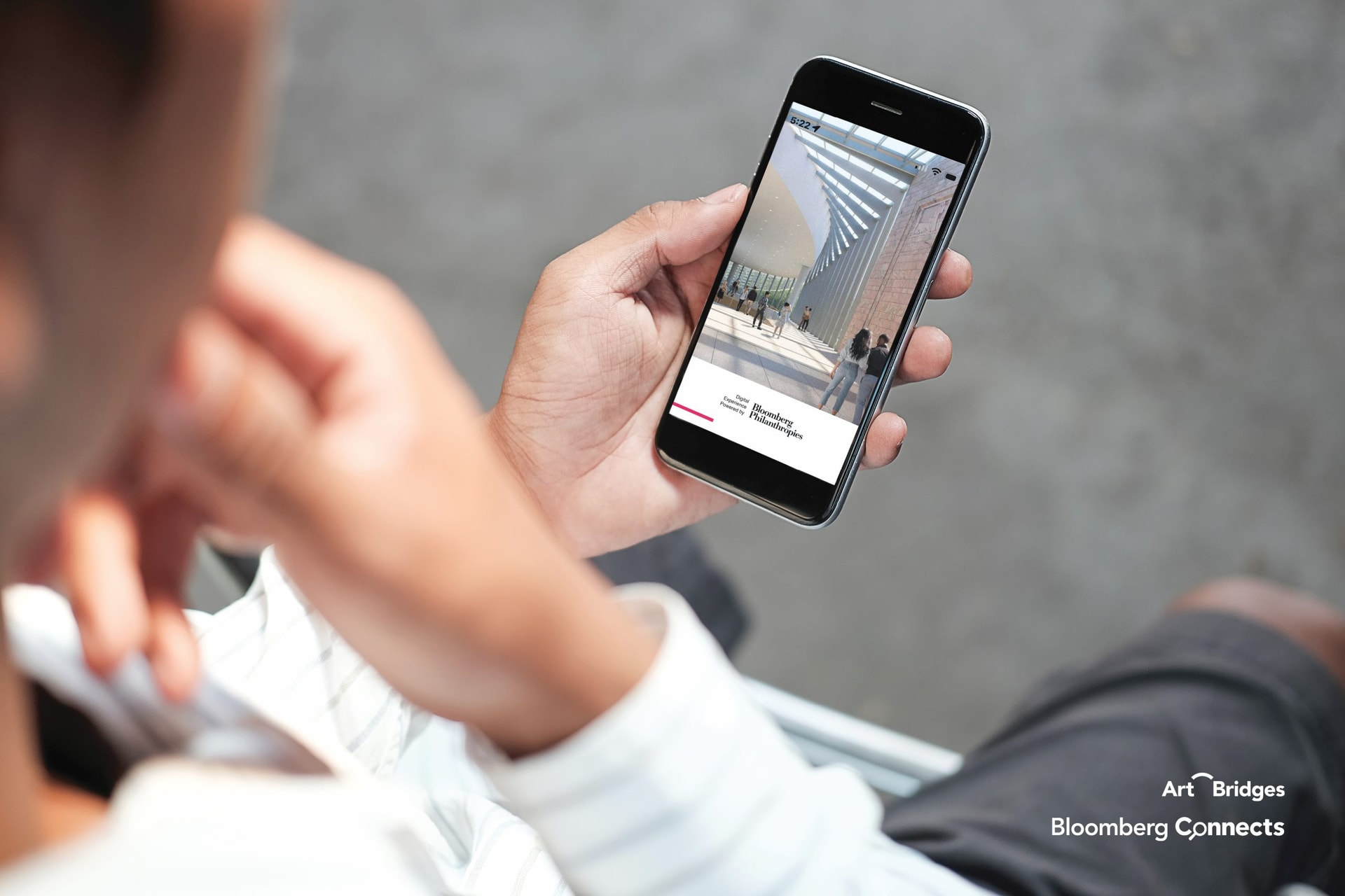 A person holds a smartphone displaying an app with the Art Bridges and Bloomberg Connects logos. The app shows an image of an art exhibit with people walking in a modern gallery space. The person uses their other hand to touch their face while looking at the phone.