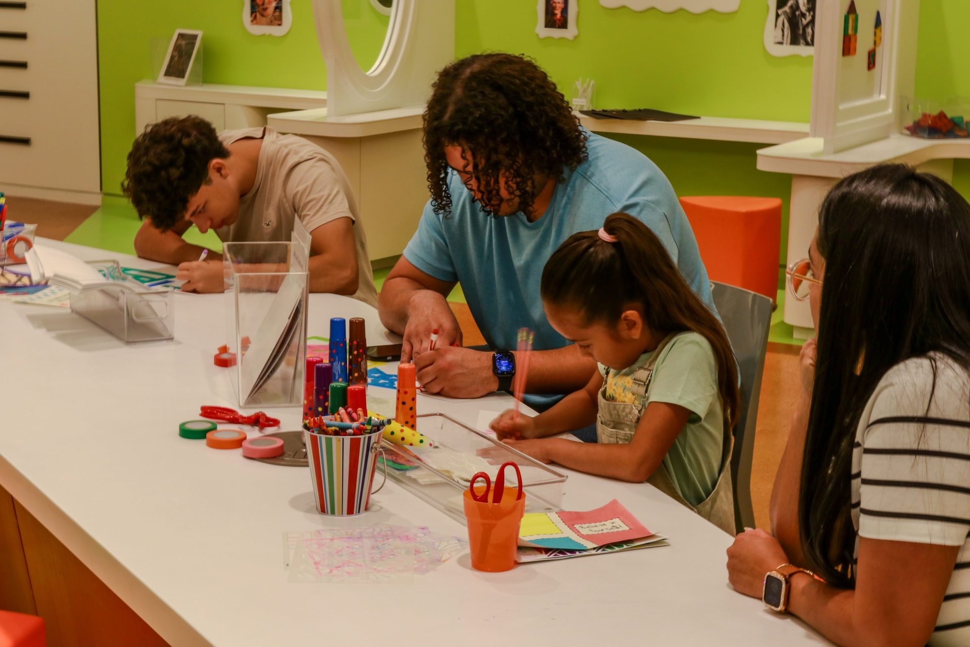 A man and a young girl are sitting at a table doing arts and crafts, with colored papers, markers, and scissors scattered on the table. Next to them, another man and a woman are also engaged in similar activities. The room has bright green walls with artwork displayed.