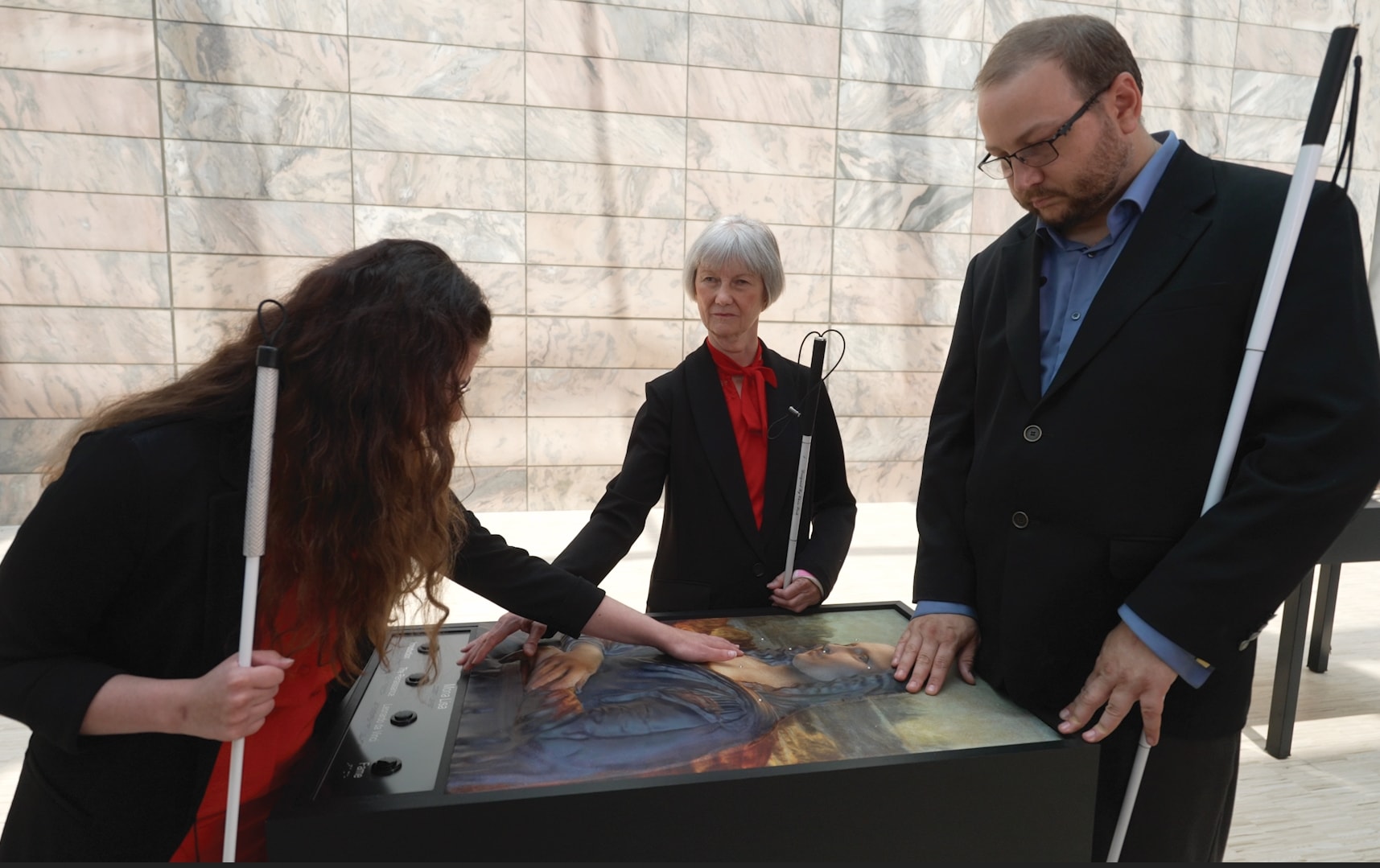 Three individuals with visual impairments explore a tactile art display with their hands. The display features textured elements for tactile interaction, and each person holds a white cane.