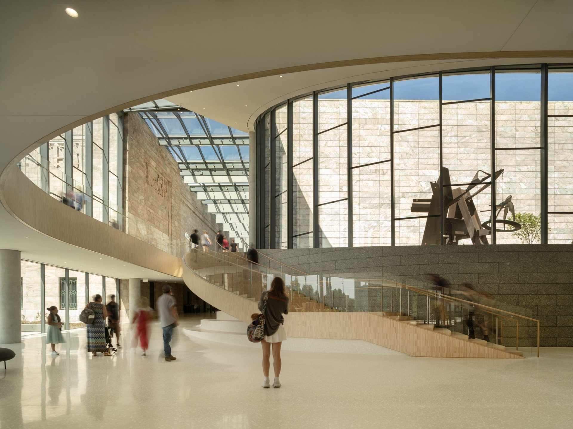 A spacious, modern interior showcases a curved staircase leading to an upper level, with people walking and mingling. Large glass windows illuminate the area, and a metallic sculpture is displayed prominently against a textured wall. Natural light fills the open space.