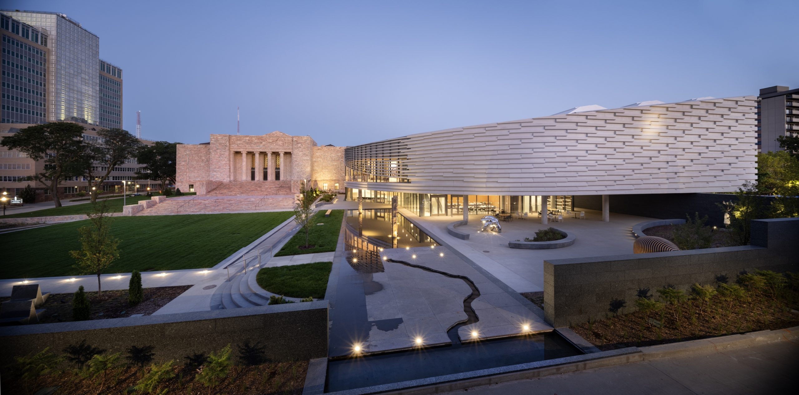 Modern art museum exterior at dusk with contemporary architecture. Features include a sleek, curving façade, expansive plaza, manicured lawn, and reflecting pool. Warm lighting highlights the building's design against a dusky sky.