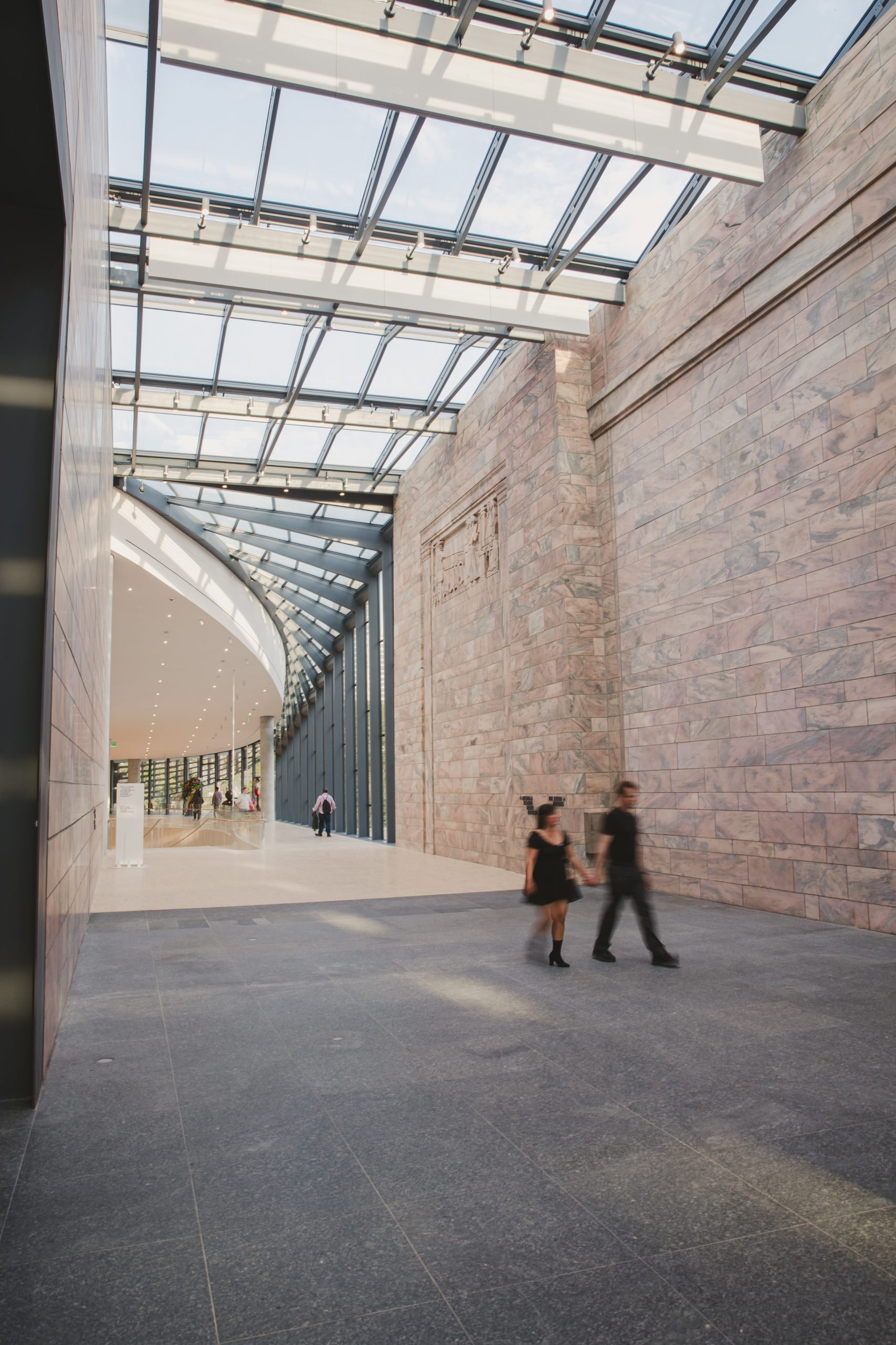 Two people walk through a modern, spacious corridor with high glass ceilings, allowing natural light to illuminate the stone walls and gray floor. The corridor leads to a brightly lit area with people in the distance.