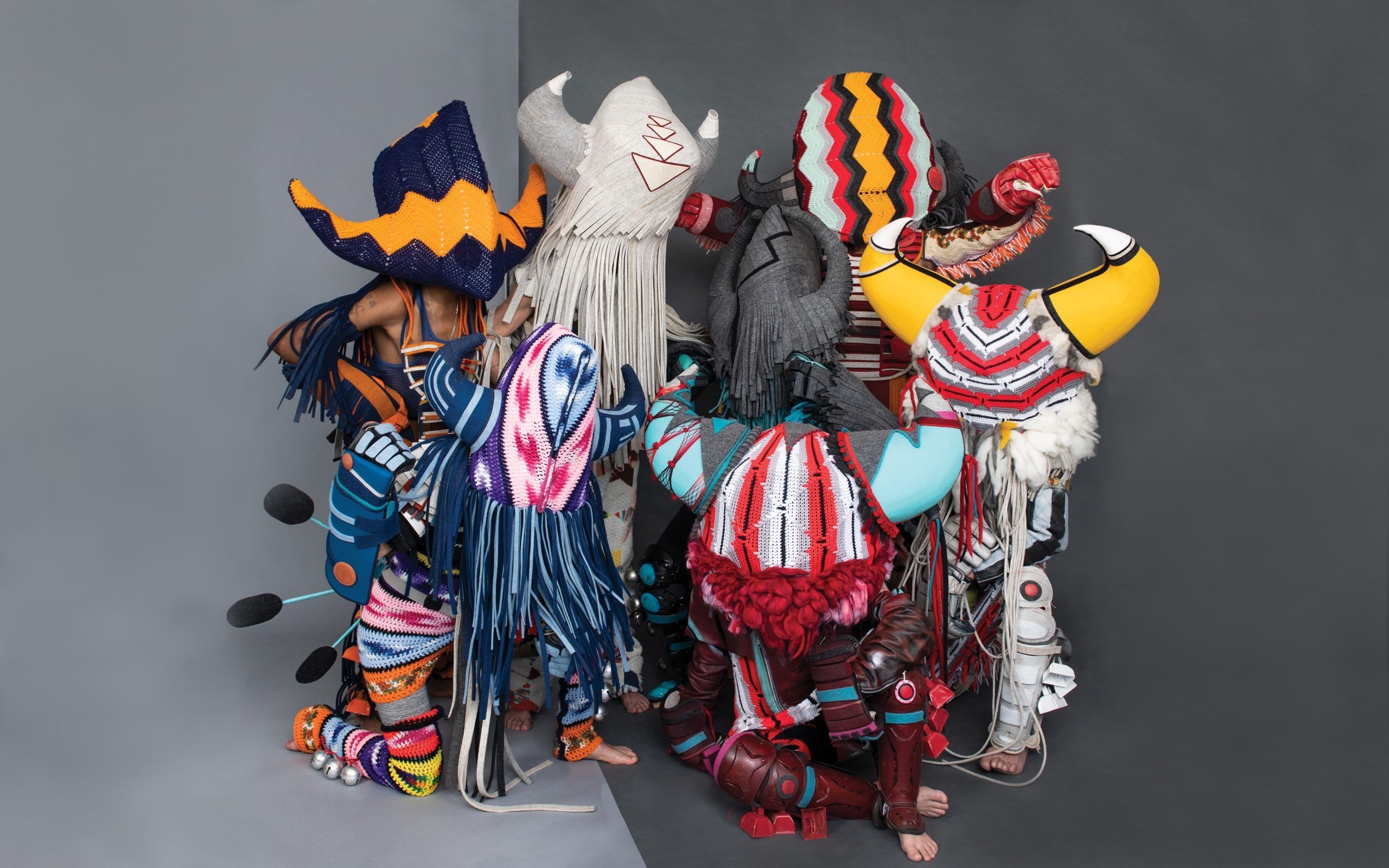 A group of people wearing vibrant, abstract costumes with large, colorful headpieces stand in a semi-circle. The costumes feature various patterns and textures, including stripes and fringe, set against a two-toned background.