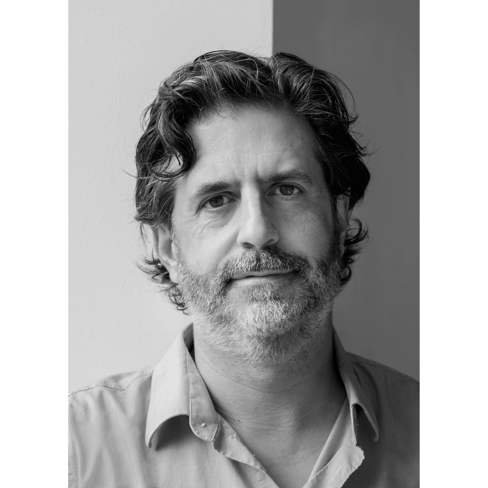 Black and white portrait of a middle-aged man with wavy hair and a beard, wearing a collared shirt. He is looking directly at the camera in front of a light-colored background.