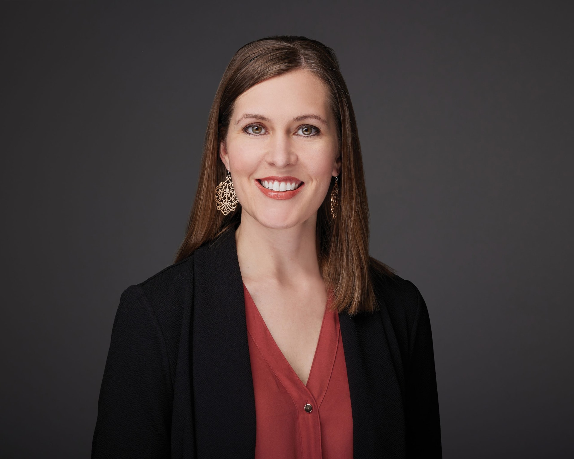 A woman with shoulder-length brown hair smiles at the camera. She is wearing a black blazer, a rust-colored blouse, and large, intricate gold earrings. The background is a plain dark gray.