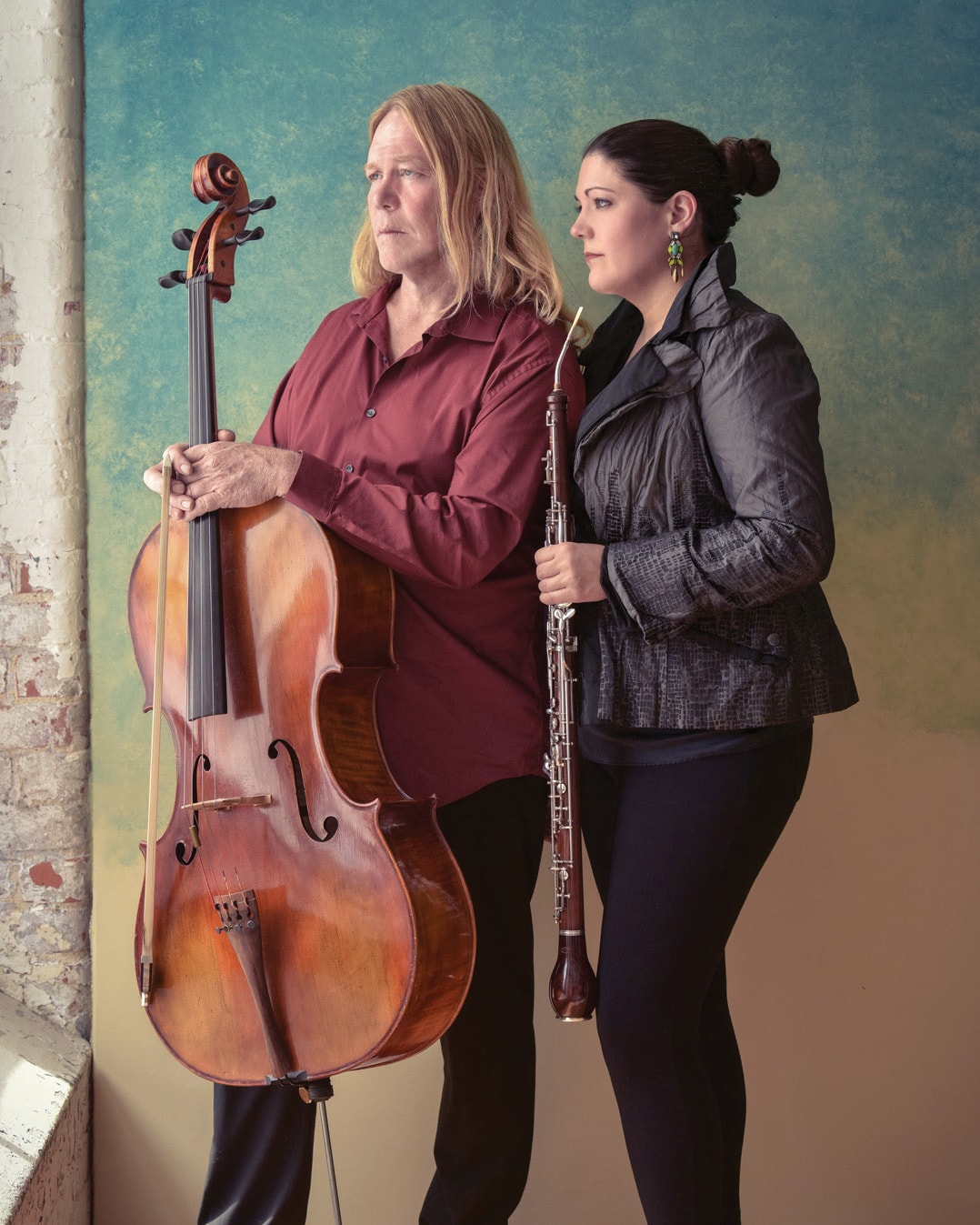 A man with long hair holds a cello, wearing a red shirt, while standing next to a woman with dark hair holding an oboe, wearing a gray jacket. They stand against a textured wall with a mix of green and blue colors.