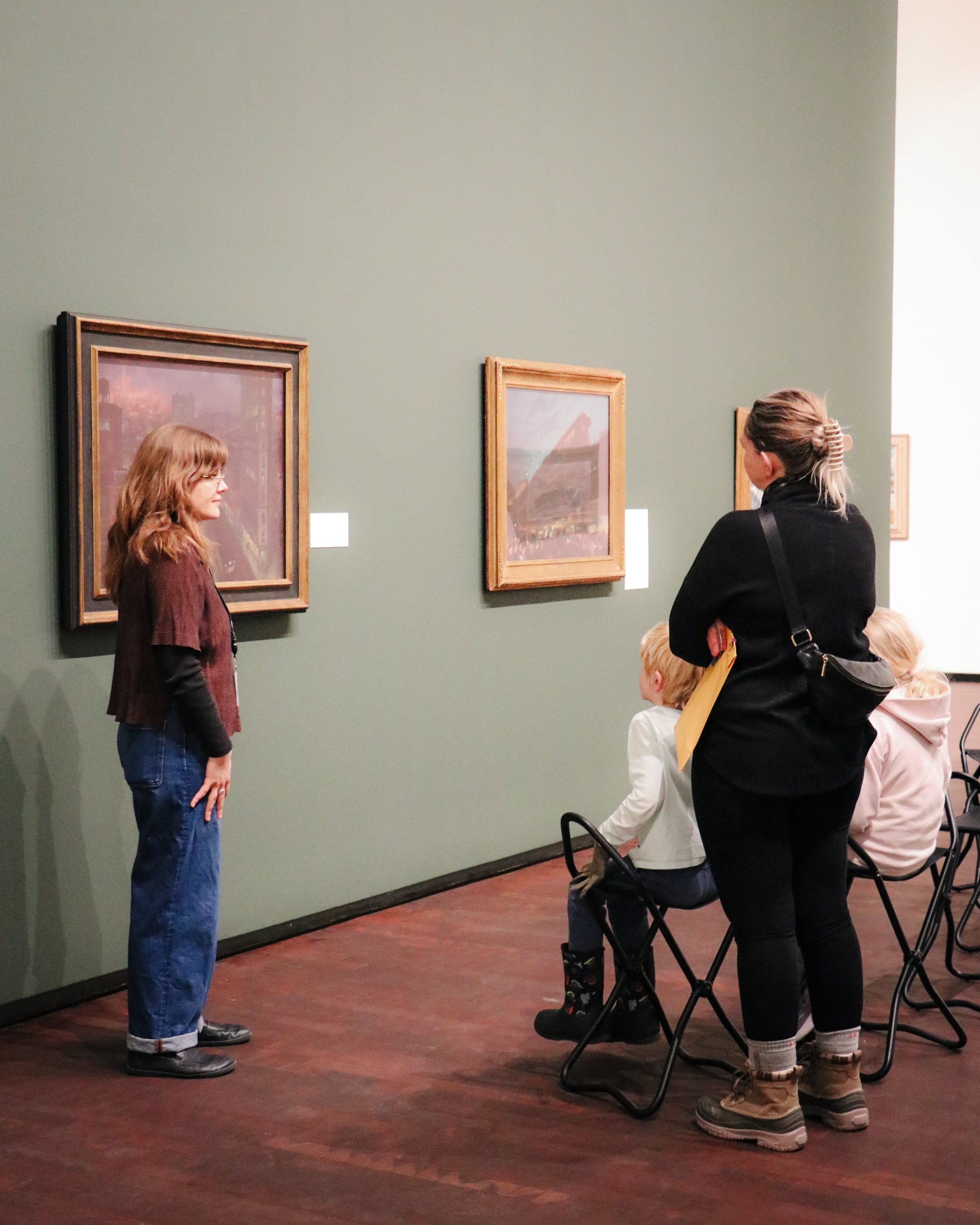 A woman and two children sit on folding chairs, facing another woman who stands next to a painting on a green wall in a gallery. The scene suggests they are discussing the artwork.