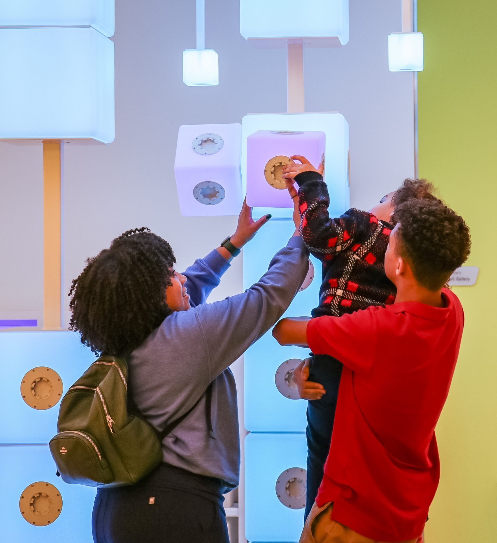 A family interacts with a light exhibit. A woman with a backpack helps a young child adjust a glowing white cube with embedded gears, as a man in a red shirt supports the child. The background features similarly glowing cubes.