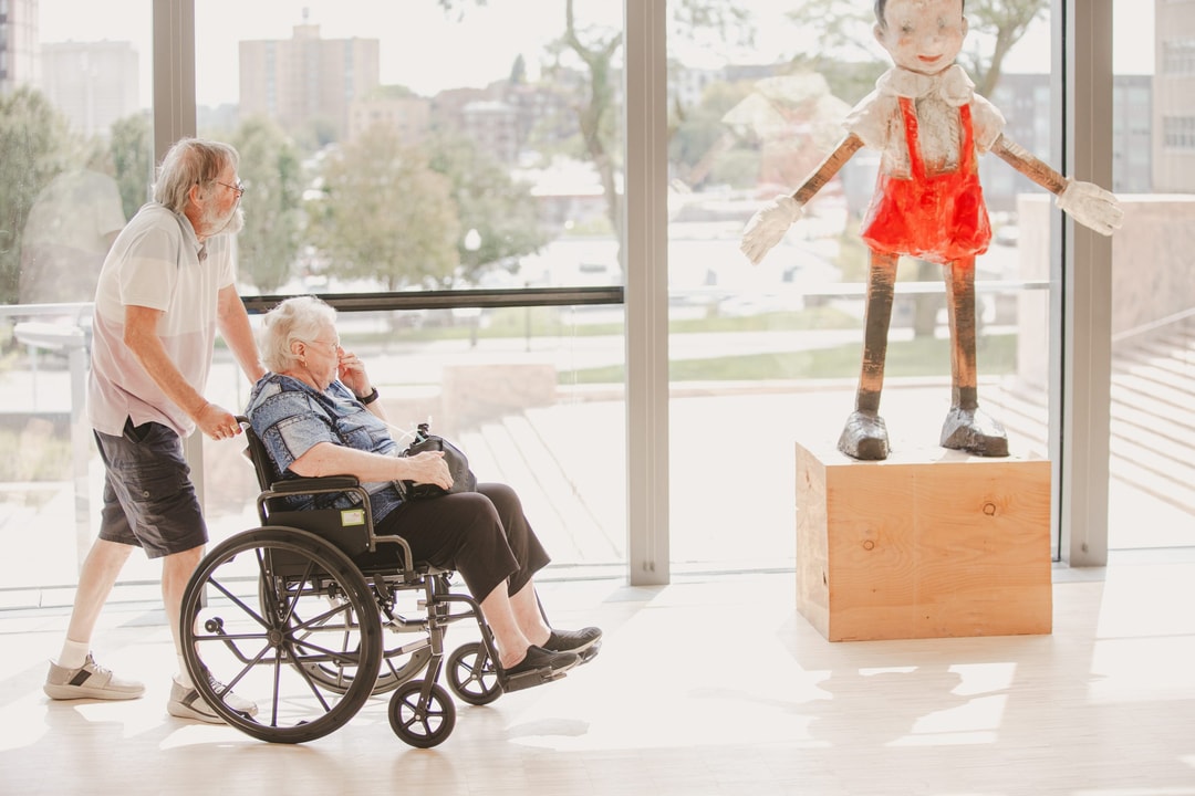 An elderly man pushes a woman in a wheelchair past a large Pinocchio sculpture in a bright, modern art gallery. The room has large windows with a cityscape view in the background.